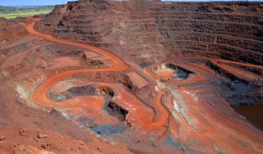 Looking down on a large open cut iron ore mine in Australiaaas remote Pilbara region.  At the bottom of the pit at the end of the red winding road a dump truck is being loaded with ore.  Other mine vehicles appear tiny in the huge pit.  Beyond the open cut are green hills.  Taken with permission during a guided tour of the mine.