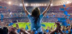 On the foreground a group of cheering fans watch a sport championship on stadium. One girl stands with his hands up to the sky. People are dressed in blue colors. A long-range shot of a stadium field, floodlights and seating. A green field, with painted white lines, is visible in the foreground. In the background are diffuse out-of-focus stadium seats. Large, bright floodlights are in the top-left and top-right corners of the image.