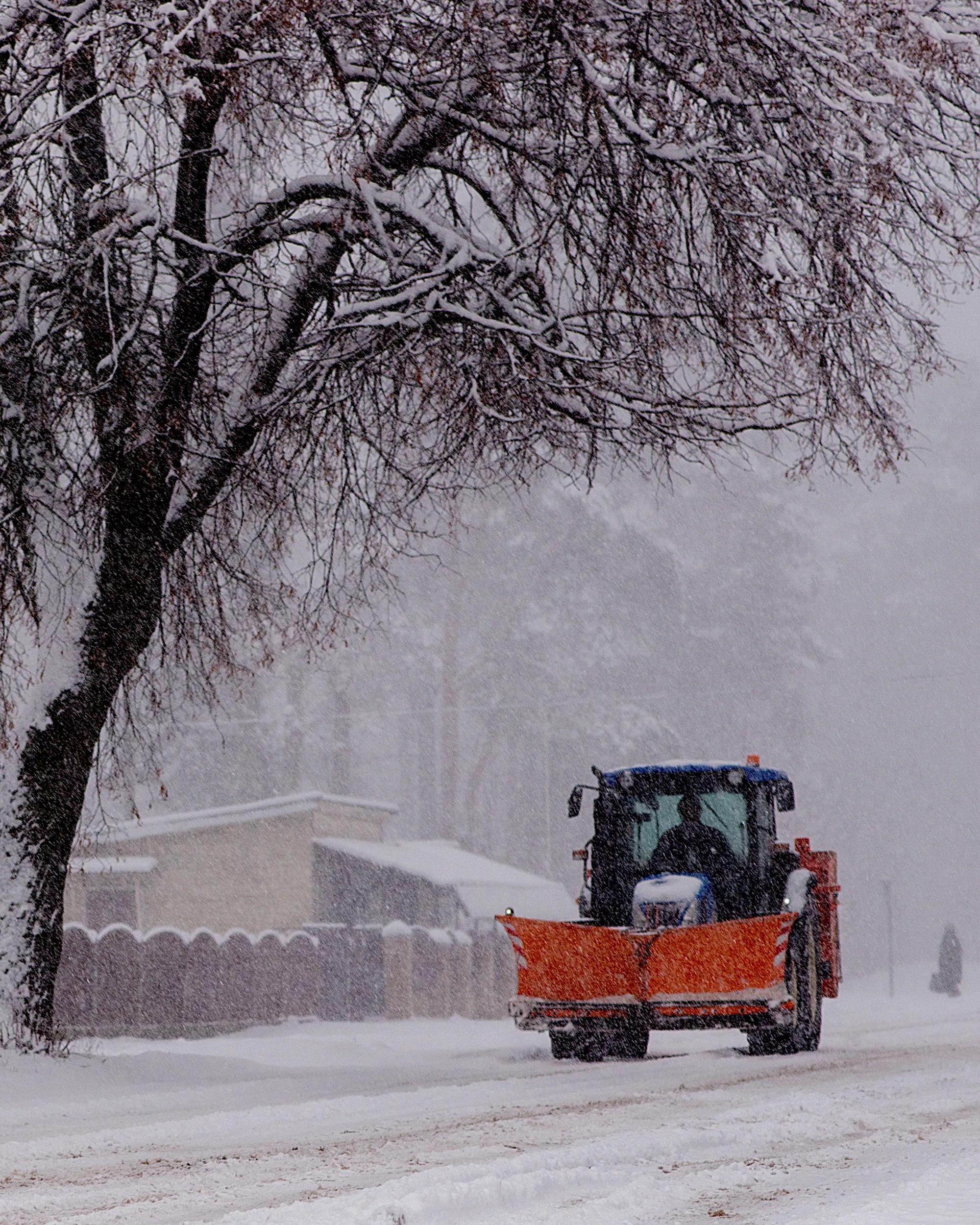 A front view of a snow plow pushing through heavy snowfall, with the driver visible inside.