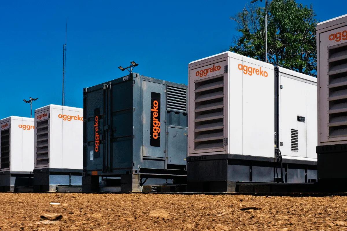 A row of Aggreko generator units on a gravel surface under a clear blue sky.