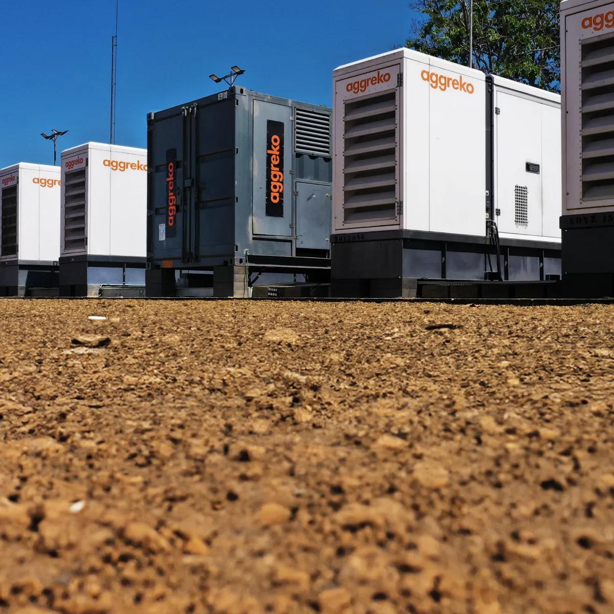 A line of large industrial generators against a blue sky and a flat, barren landscape.