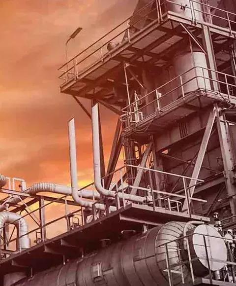 A close-up, low-angle shot of a large, gray industrial structure with pipes and walkways against an orange sky.