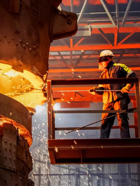 A close-up shot of a foundry operation where extremely hot, bright orange molten metal is pouring from a massive ladle into a container. The scene is dark, lit only by the incandescent glow of the metal, and sparks are flying.