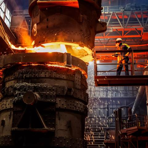 A close-up shot of a foundry operation where extremely hot, bright orange molten metal is pouring from a massive ladle into a container. The scene is dark, lit only by the incandescent glow of the metal, and sparks are flying.
