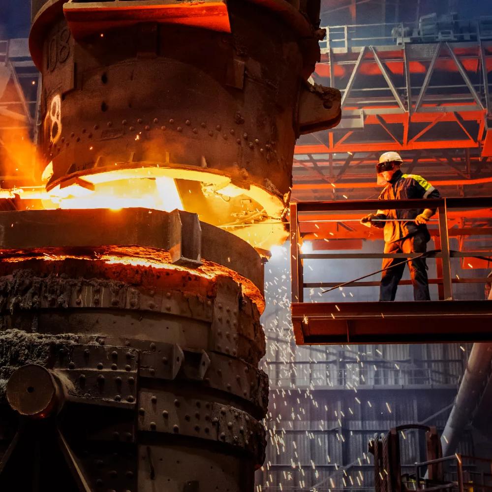 A close-up shot of a foundry operation where extremely hot, bright orange molten metal is pouring from a massive ladle into a container. The scene is dark, lit only by the incandescent glow of the metal, and sparks are flying.