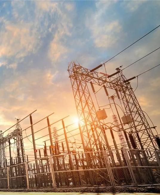 A complex electrical substation with transmission lines and equipment silhouetted against a bright sunset.