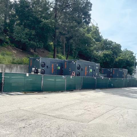 A white Aggreko pickup truck, parked near a temporary green fence. Behind the fence, dark, containerized industrial units are visible, positioned against a concrete retaining wall and green hill.