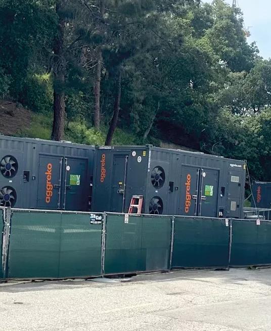 A white Aggreko pickup truck, parked near a temporary green fence. Behind the fence, dark, containerized industrial units are visible, positioned against a concrete retaining wall and green hill.