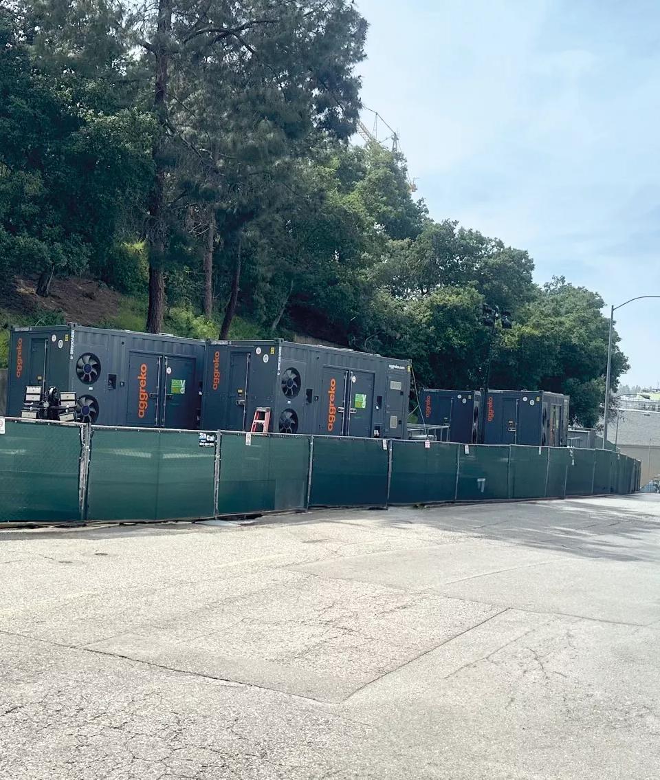 A white Aggreko pickup truck, parked near a temporary green fence. Behind the fence, dark, containerized industrial units are visible, positioned against a concrete retaining wall and green hill.