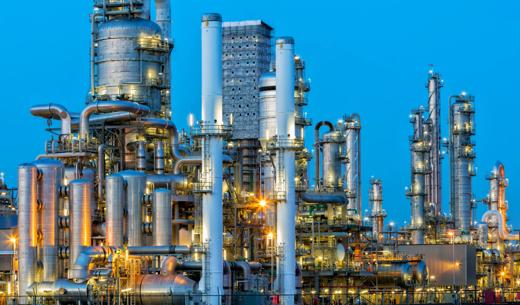 Industrial complex with numerous tall pipes and structures, illuminated against a blue evening sky.