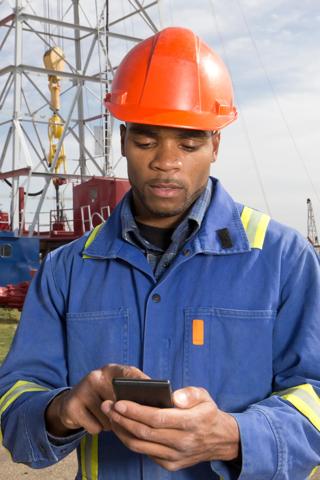 A royalty free image of an oil worker using a smart phone to text while at an oil rig.