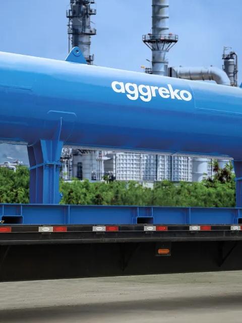 Échangeur de chaleur industriel Aggreko transporté sur un camion à plateau, avec une installation industrielle en arrière-plan.