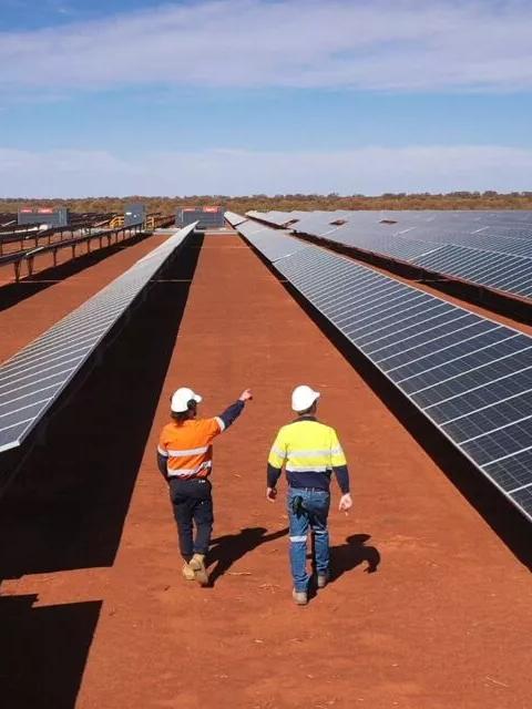An aerial view of vast, closely-packed rows of solar panels extending across a red, desert-like landscape. Two workers in hard hats and safety vests are looking out over the panels.
