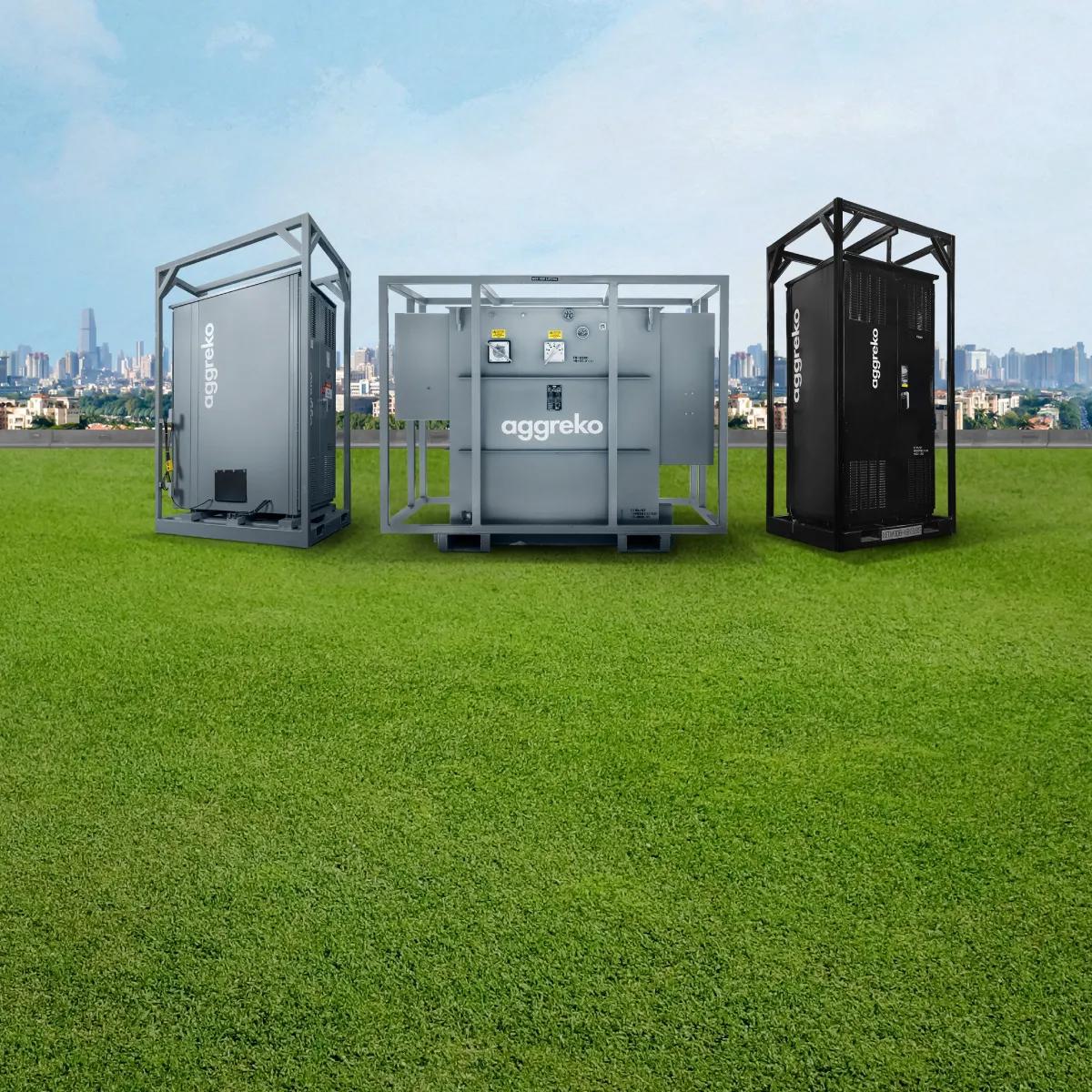 A grey transformer, a grey switchgear system and a black distribution panel, all with the Aggreko logo, standing on a green field with a city's skyline in the background