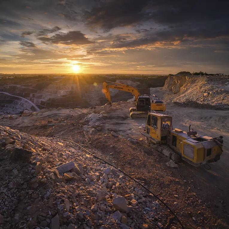 A mining site at sunset, with yellow excavators near a rocky hillside.
