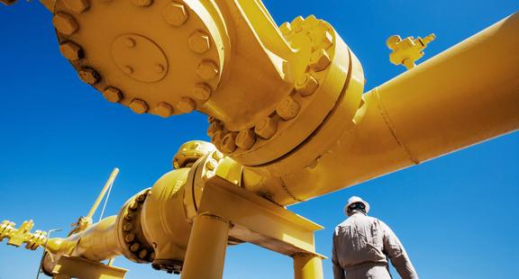 Worker in protective gear standing next to large yellow industrial gas pipes under a clear blue sky