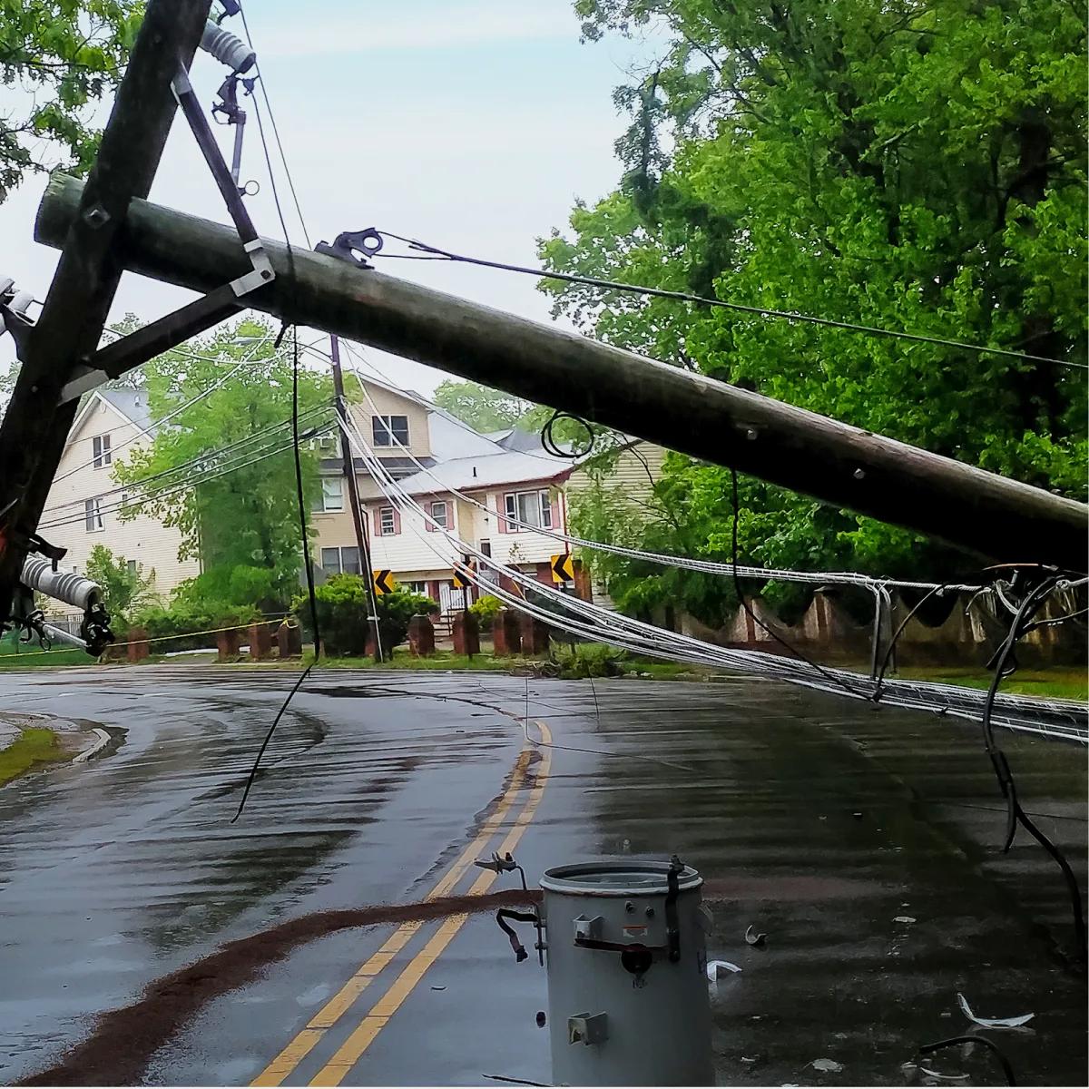 A downed utility pole with wires on a wet road, surrounded by greenery and a house in the background.