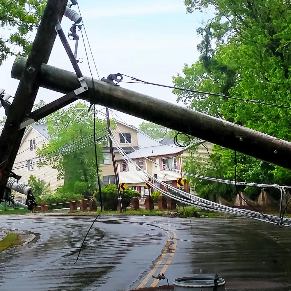 A downed utility pole with wires on a wet road, surrounded by greenery and a house in the background.
