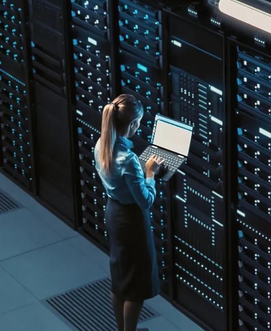 A high-angle, wide shot inside a vast data center showing countless rows of tall, dark server racks. A woman stands in the foreground, facing the racks while holding a laptop and troubleshooting.