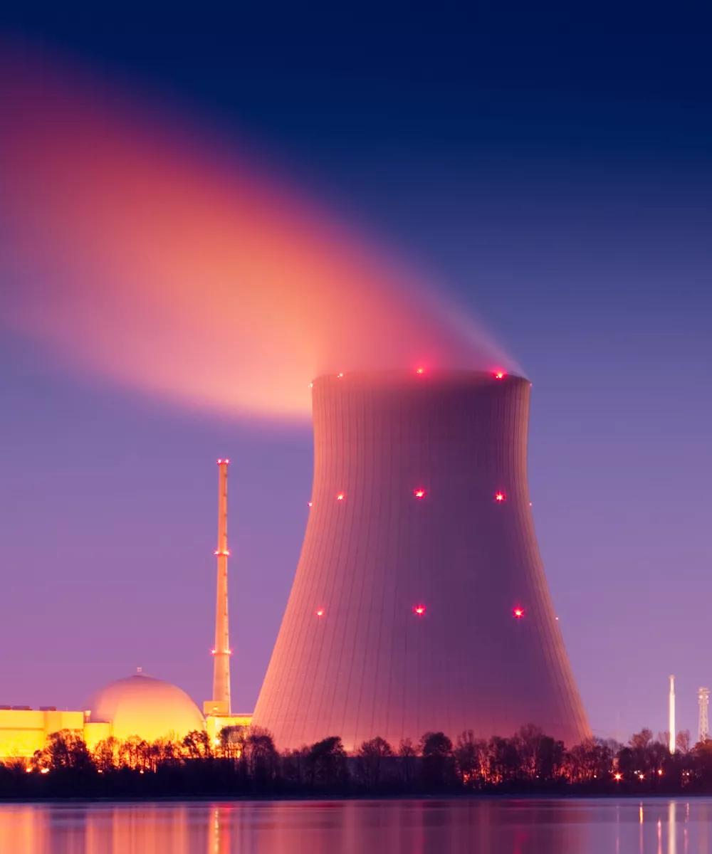 A nuclear power plant with a glowing tower and a in front of water at dusk