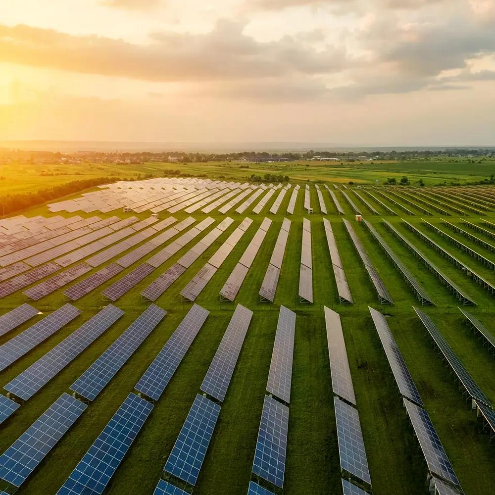An aerial view of a large utility-scale solar project with extensive rows of solar panels covering the left half of the image in a dry, open field. On the right, a complex electrical substation with large metal structures and transformers, with a white truck parked inside the fenced perimeter.