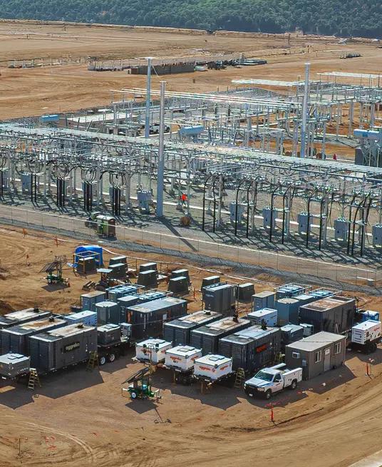 Aerial view of a variety of containerized industrial equipment and generators staged on a gravel lot near a green field with several large wind turbines in the background.