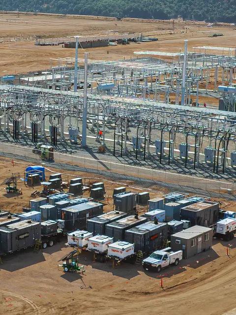 Aerial view of a variety of containerized industrial equipment and generators staged on a gravel lot near a green field with several large wind turbines in the background.