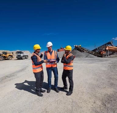 Open- pit mine engineer and workers wearing protective clothes and helmets standing in front of pile of gravel and excavator. Manager holding clipboard and discussing with team. Wide angle view.