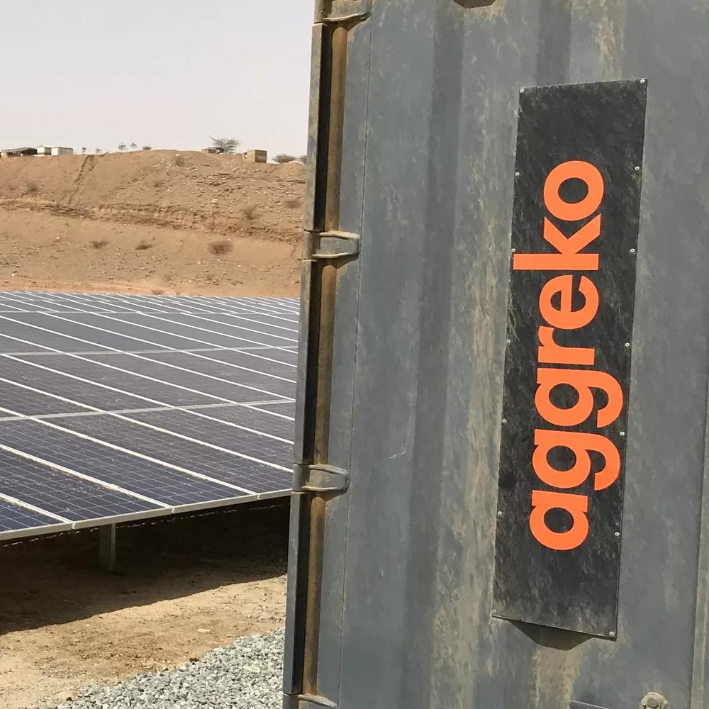 A large field of solar panels and a dark Aggreko unit on a gravelly surface.