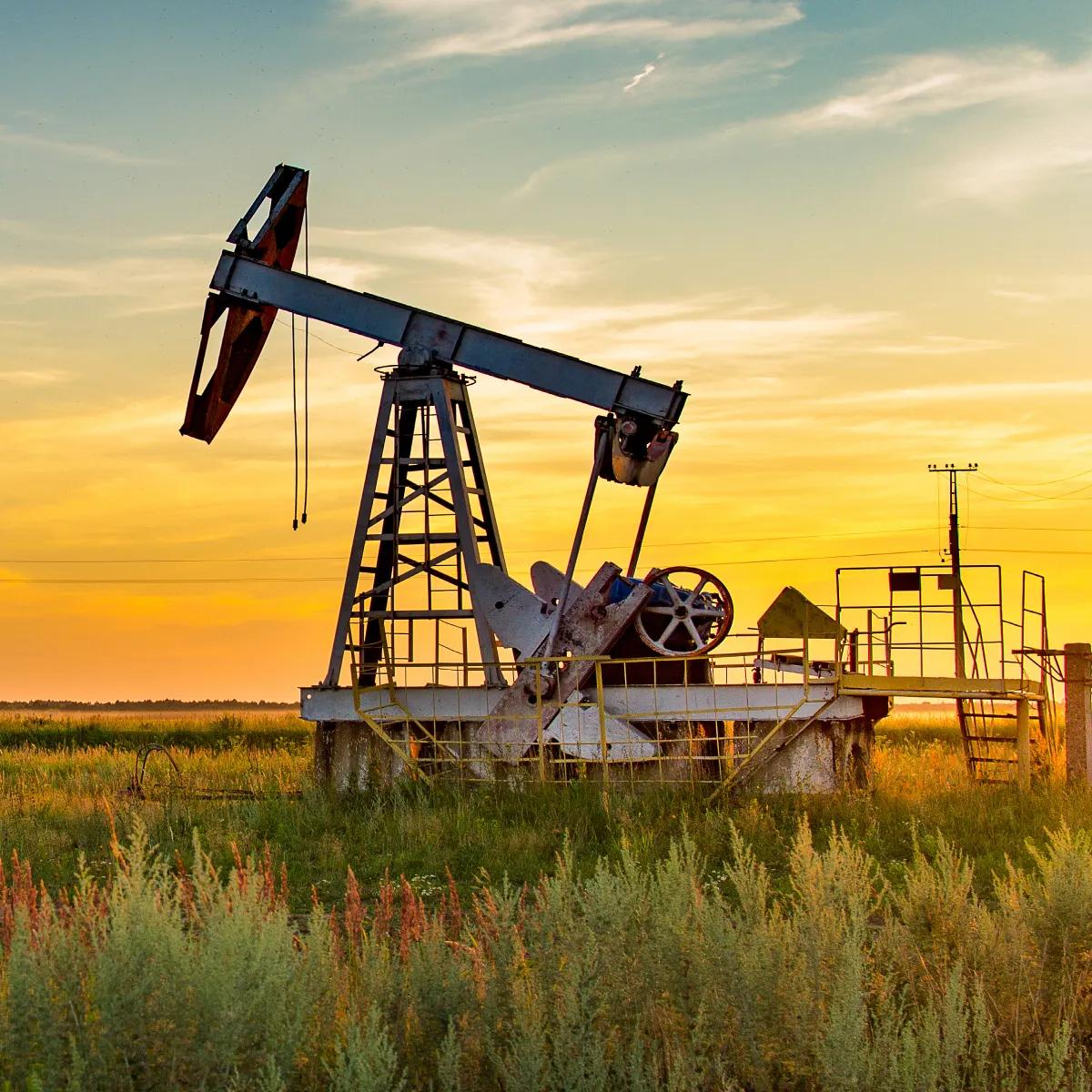 An oil pump jack stands against a backdrop of rolling green hills and distant mountains under a clear sky.