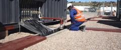 A worker in a safety helmet and orange vest kneels by equipment, handling cables near a generator in a gravelly area.