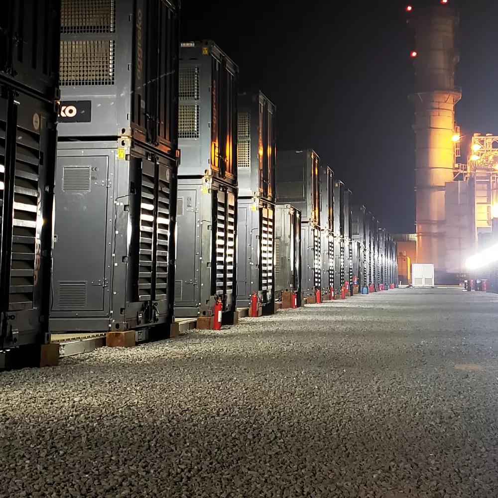 A long, perspective view of a row of dark, containerized Aggreko industrial units at night, brightly illuminated by ground-level spotlights. Industrial structures and a glow from flares or light towers are visible in the distance.