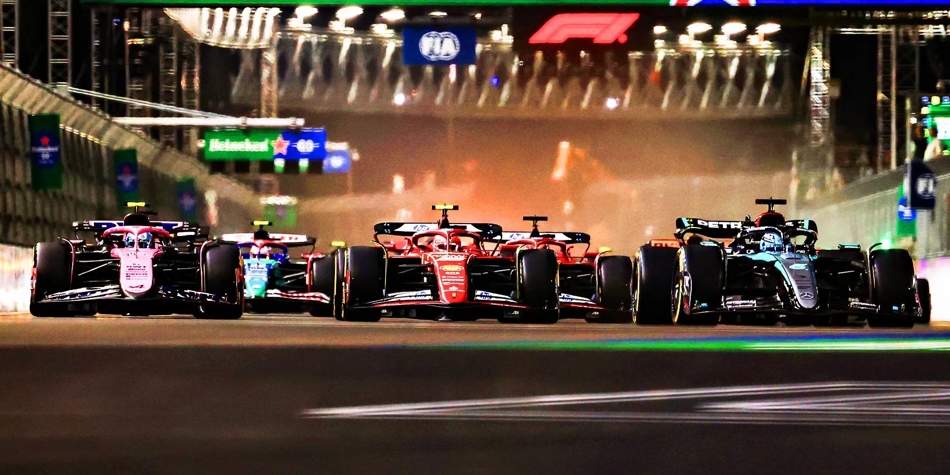 A wide, low-angle shot of a group of F1 race cars speeding down a racetrack at night, with their headlights shining and the track illuminated by stadium lights.