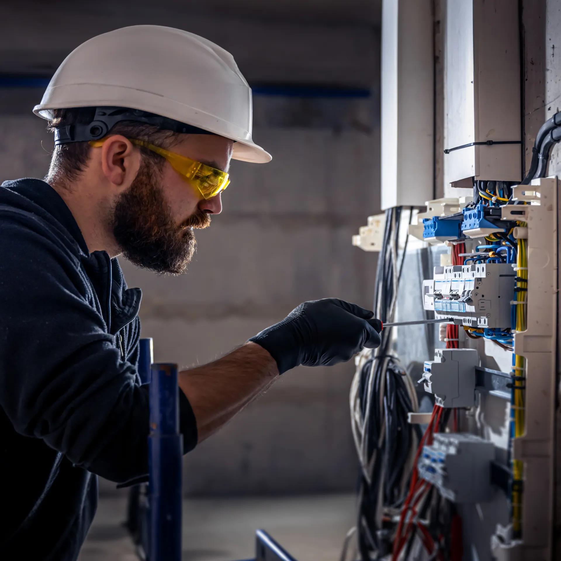 A focused worker in safety gear repairs electrical equipment on a wall in a construction setting.