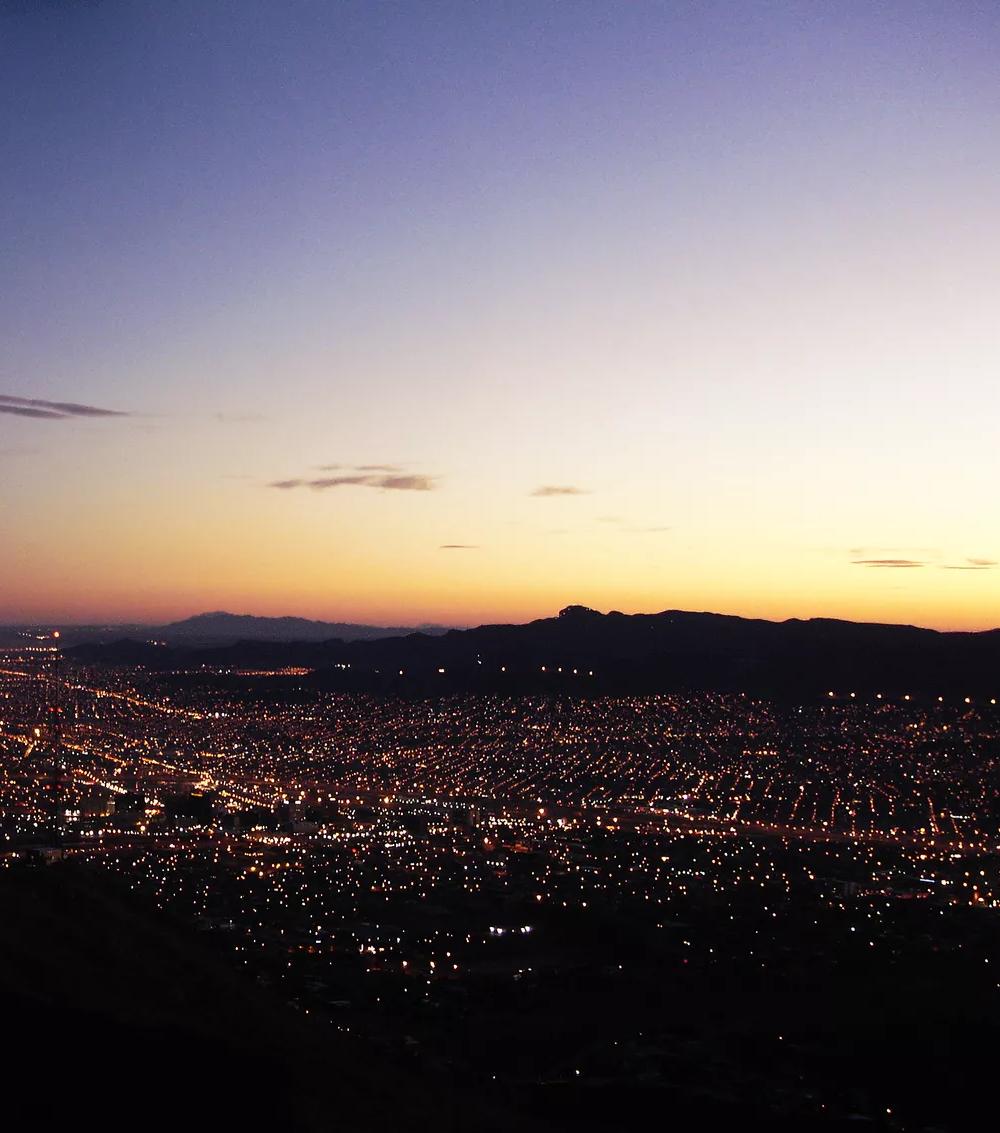 Vue panoramique d’une ville au crépuscule, avec de nombreuses lampes qui scintillent et un coucher du soleil derrière des montagnes lointaines.