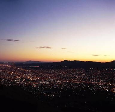 A panoramic view of a city at dusk, with numerous lights twinkling as the sun sets behind distant mountains.