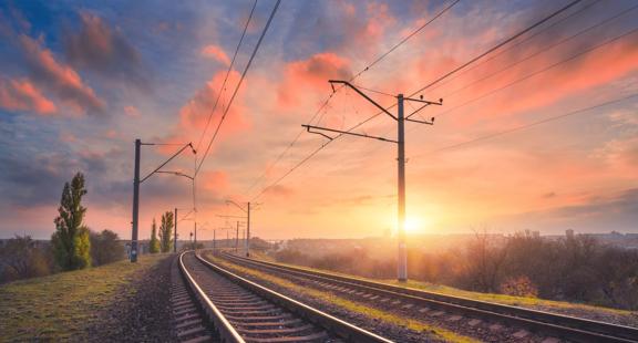 View of a railroad surrounded by power lines against an orange sunset in a green field