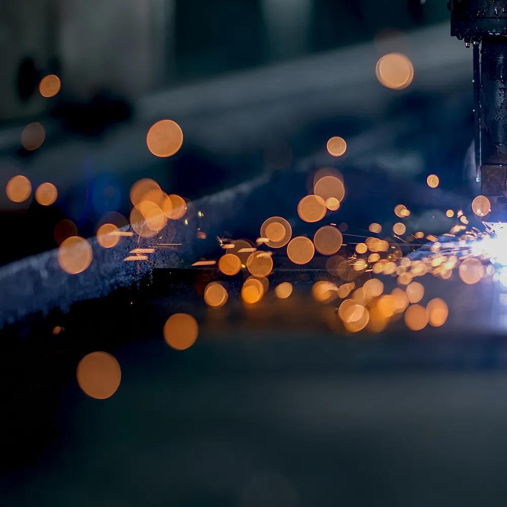 A close-up, dark image of a welding operation, showing a shower of bright orange sparks against a blurry industrial background.