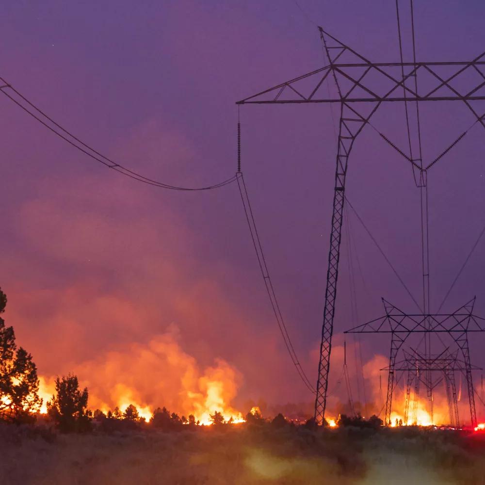 Silhouettes of power lines against a dark sky, with flames and smoke rising from a wildfire in the background.