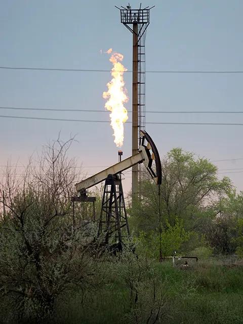 An oil rig with a flame burning at the top, surrounded by greenery and a cloudy sky.