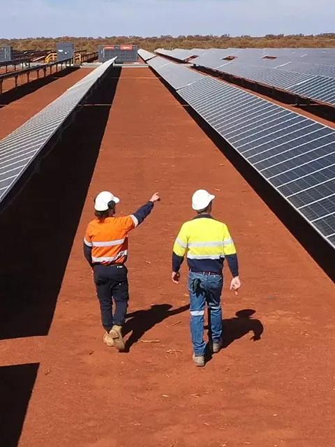 Aerial view of 2 workers in uniform walking by multiple rows of solar panels. 
