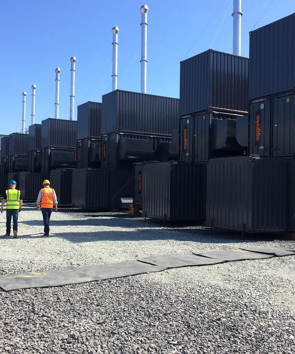 Several rows og black Aggreko industrial units stacked on one another, with 3 workers in uniform walking by.
