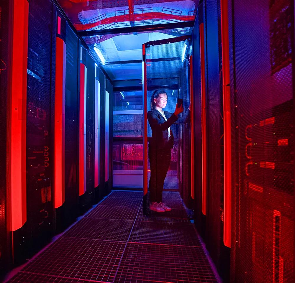 A dark, stylized shot down a server room aisle, dominated by vertical strips of alternating intense red and cool blue light. A female technician in business attire stands on the right side of the aisle, looking at a device (possibly a tablet or screen) on one of the server racks.