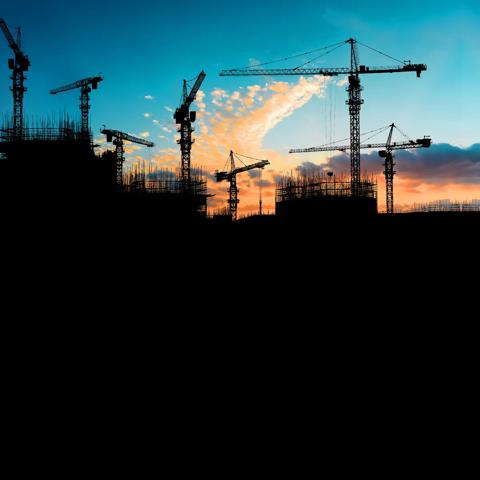 Les silhouettes de deux grues de chantier et d’une ossature de bâtiment se détachent sur un ciel bleu foncé au coucher du soleil.