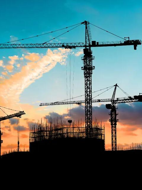 The silhouettes of two construction cranes and a building frame are shown against a dark blue sunset sky.