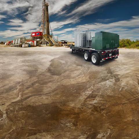 A wide image of a massive oil drilling derrick sits in the center, surrounded by industrial equipment and vehicles in a dry, open landscape under a partly cloudy blue sky. 