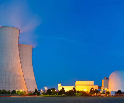 Nuclear power plant at dusk, featuring towers and a spherical containment structure, illuminated by surrounding lights.
