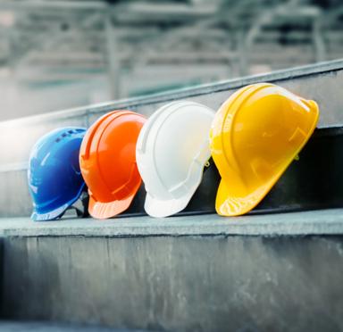 A row of four safety hats at a worksite, from left to right they're blue, orange, white and yellow