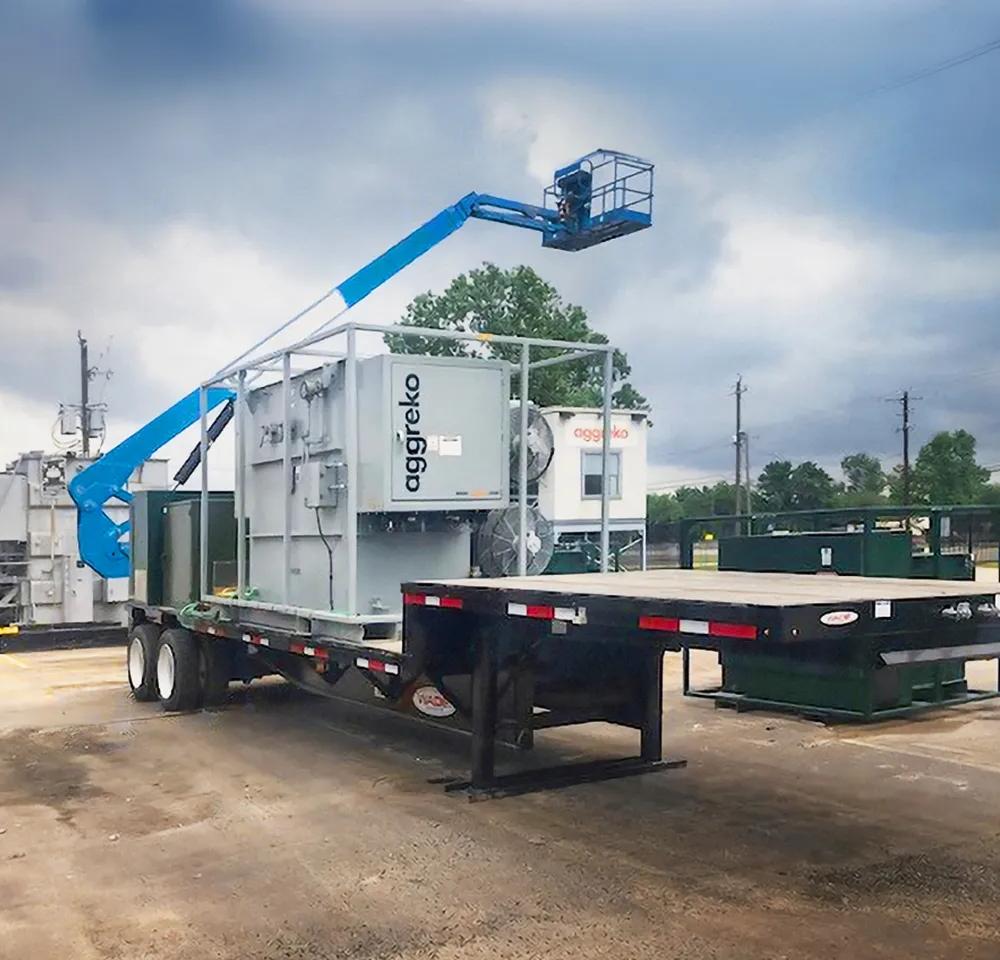 A modular, containerized Aggreko substation unit on the back of a flatbed trailer beside a green forest lining.