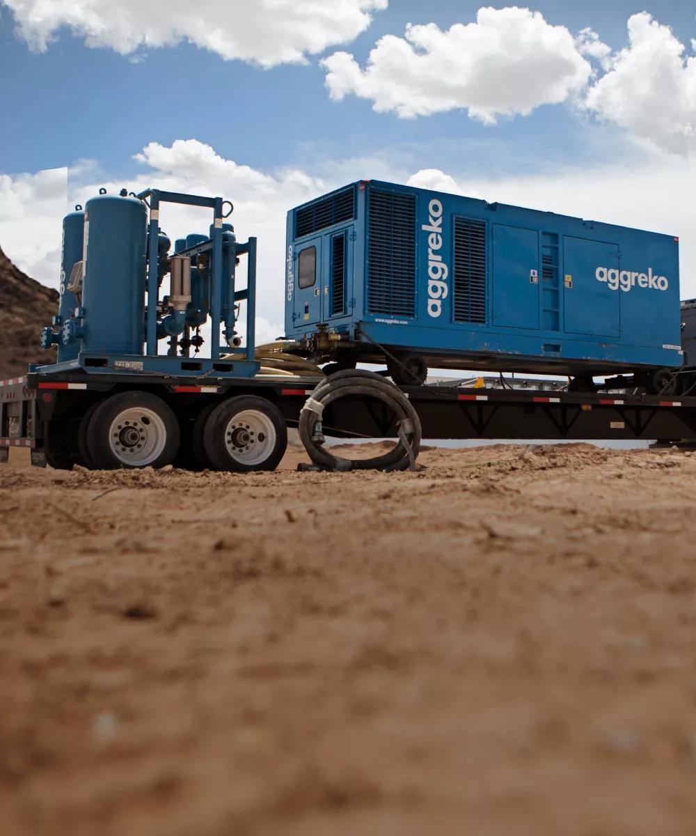 A large, dark blue containerized industrial unit is secured on a blue flatbed trailer in a dry, dirt field under a partly cloudy sky. 